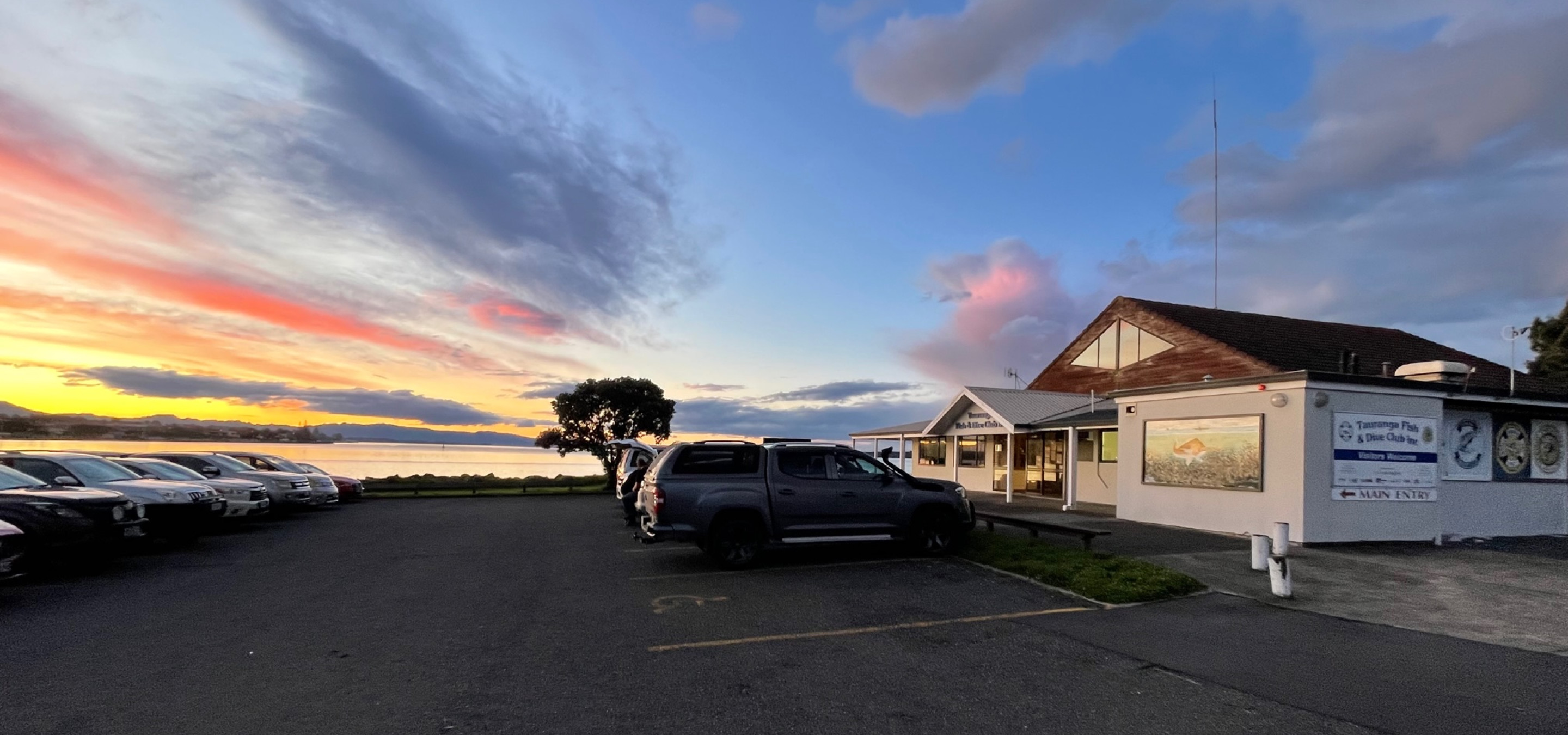 The relaxed coastal exterior of Tauranga Fish and Dive Club with a parking lot and stunning harbour sunset view.