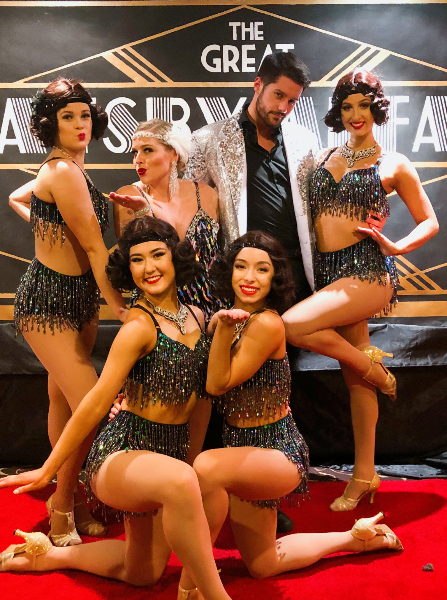 Les Follies Auckland performers, known for their glitz and glamour, pose in 1920s Gatsby-themed costumes on a red carpet.