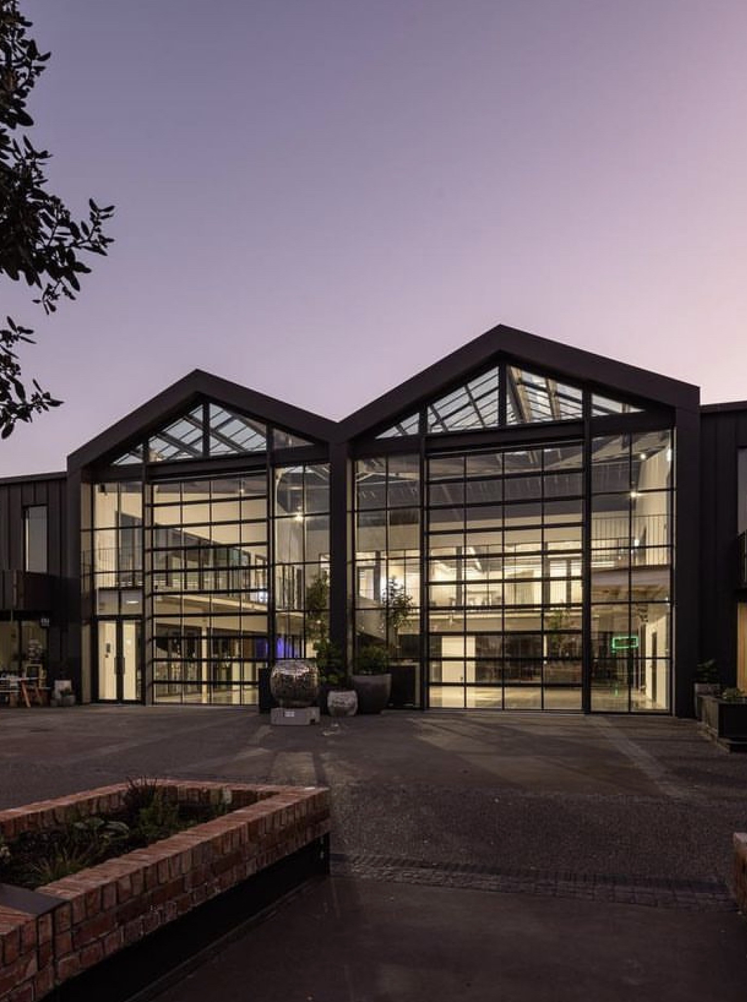 The modern industrial Atrium at Tribune in Hawke's Bay, illuminated at dusk, showcases its expansive glass exterior and courtyard.