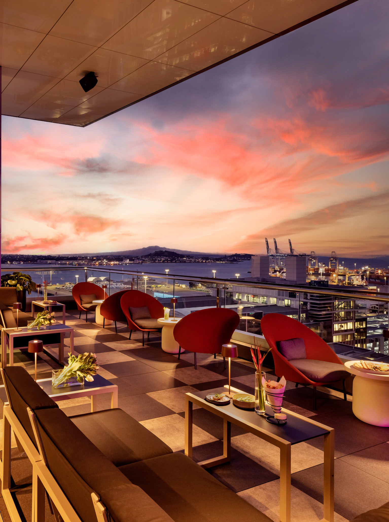 An avant-garde rooftop bar at SO/ Auckland offers playful luxury with panoramic harbor views under a vibrant sunset.
