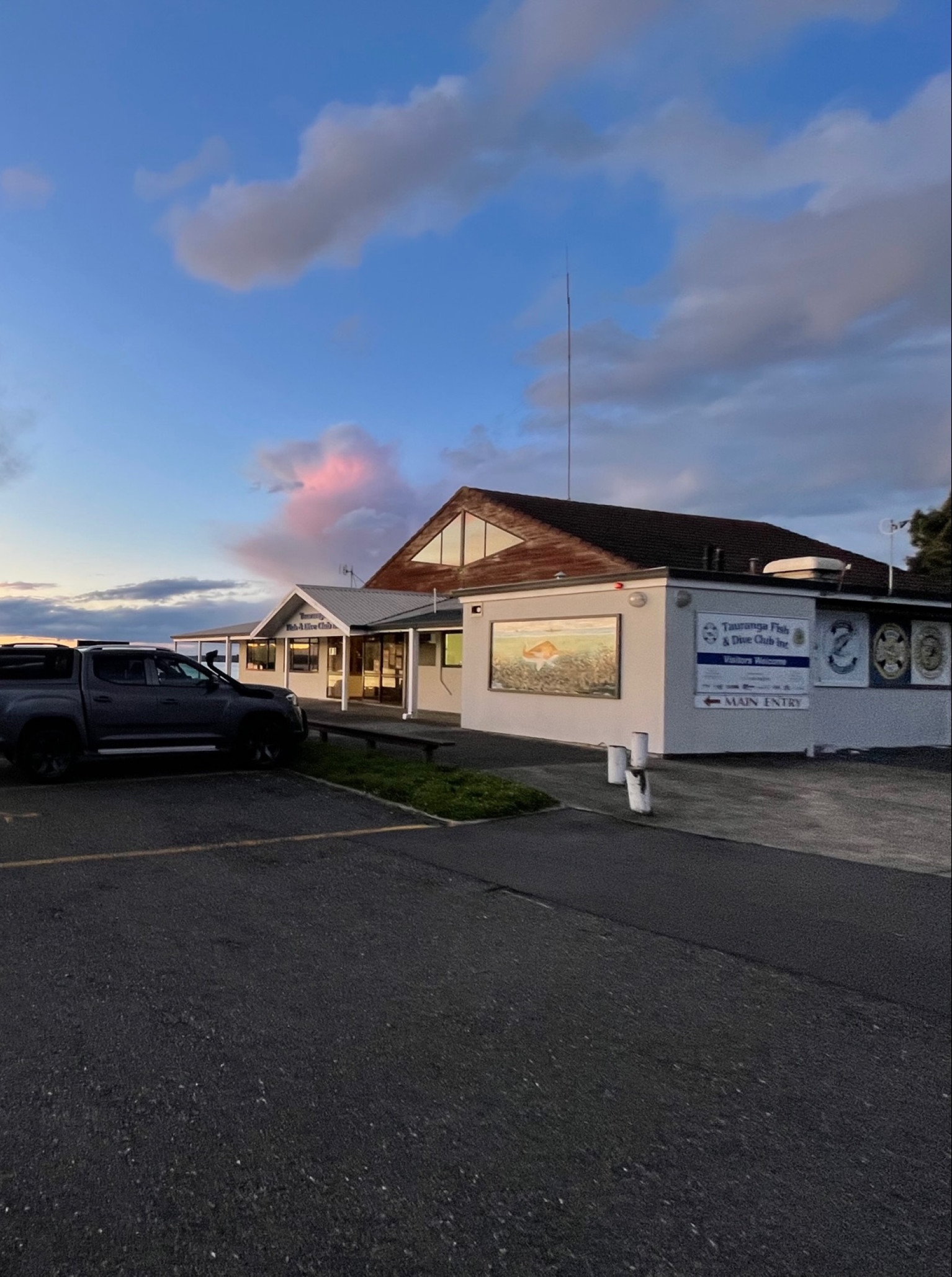 The relaxed coastal exterior of Tauranga Fish and Dive Club with a parking lot and stunning harbour sunset view.