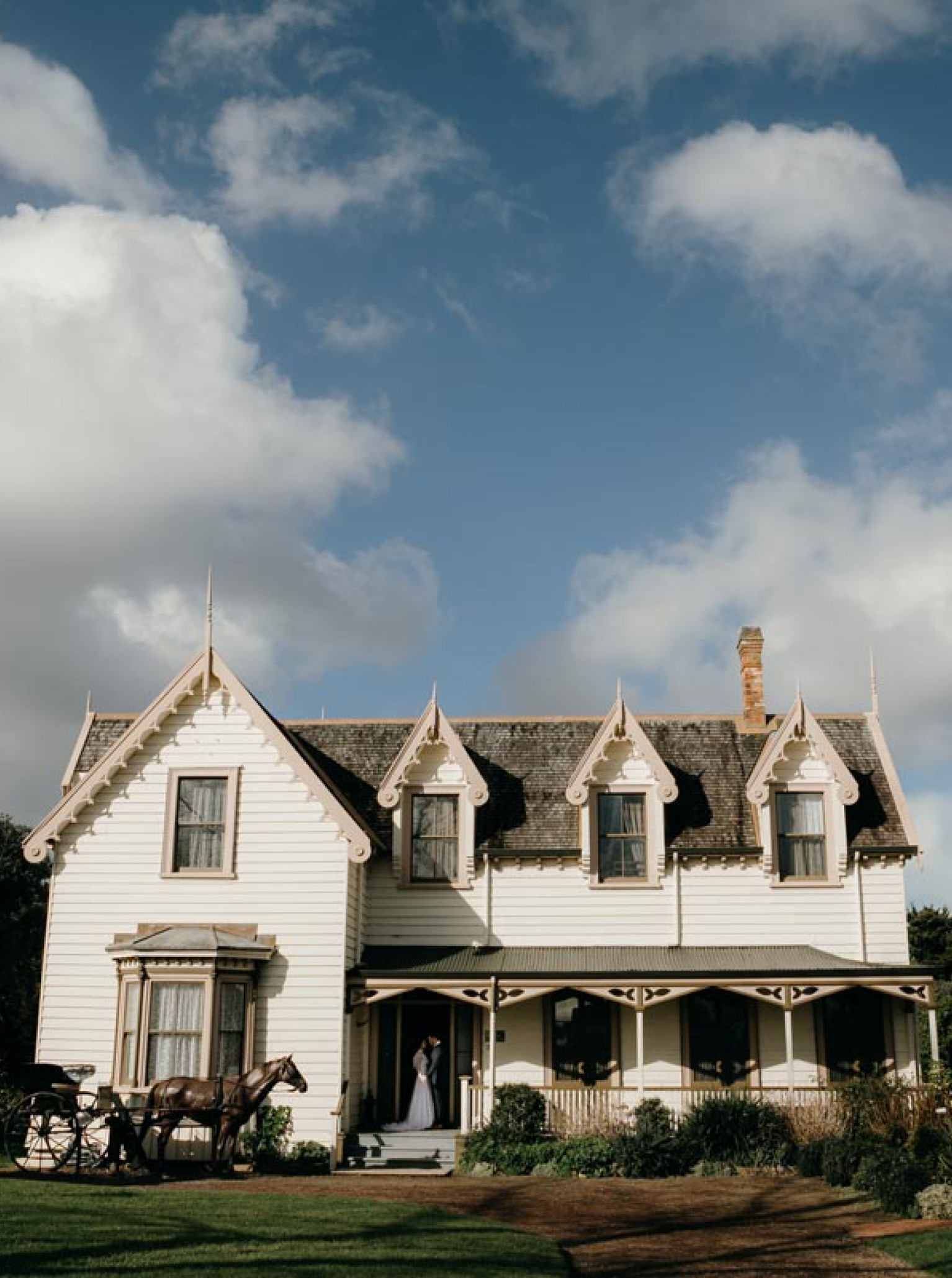 A wedding couple stands on the porch of a vintage homestead at Howick Historical Village, Auckland, with a horse-drawn carriage nearby.