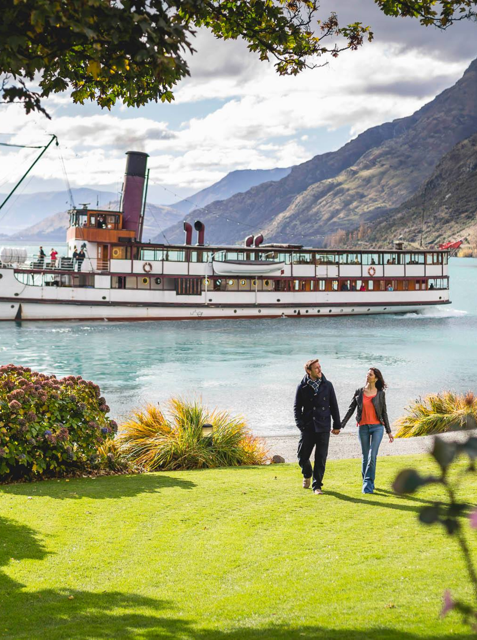 A couple strolls on the manicured lawn of Walter Peak High Country Farm, with the historic TSS Earnslaw vintage steamship cruising on Lake Wakatipu, Queenstown.