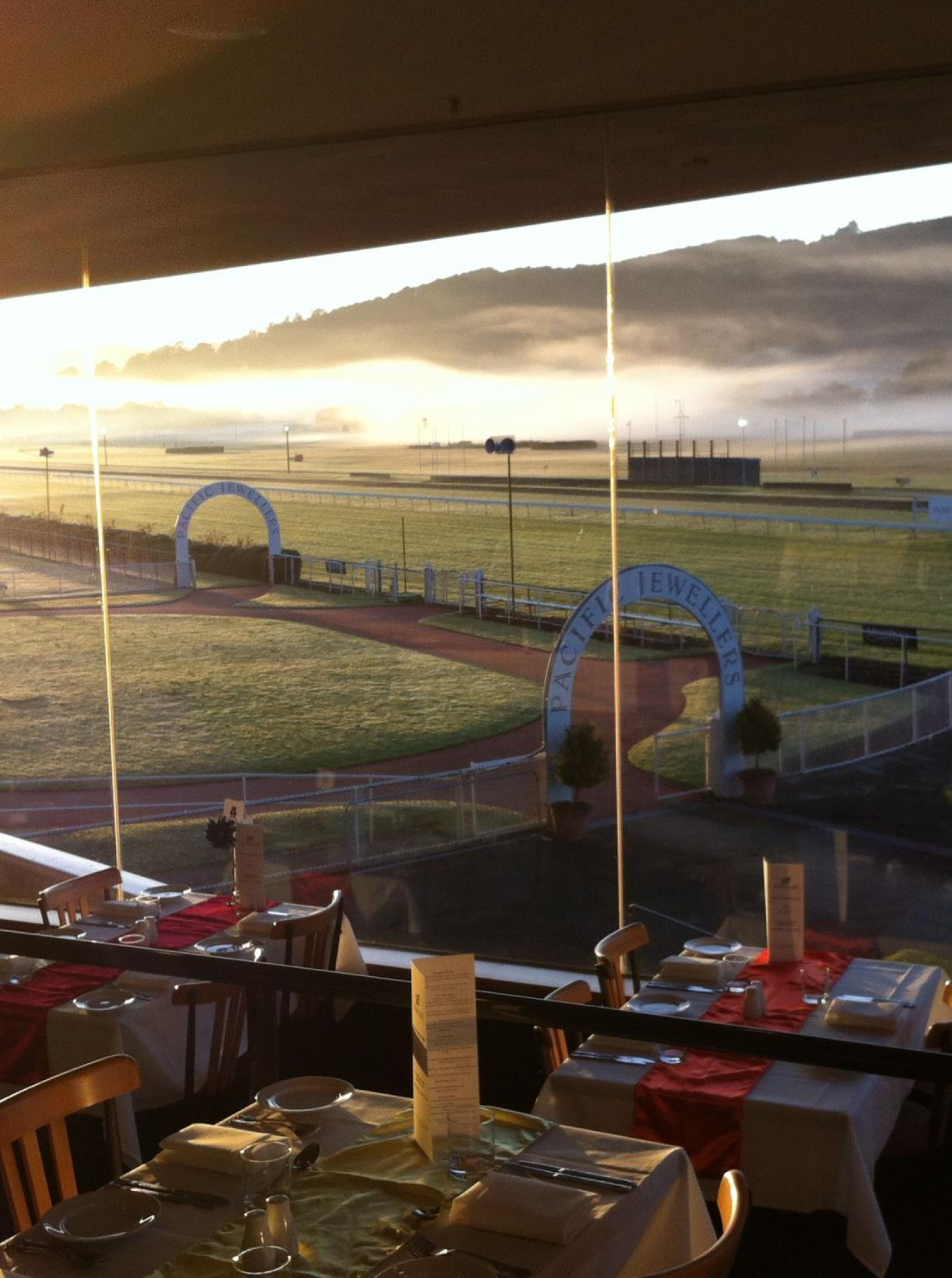 A dining room at RACE Trentham (Wellington Racing Club) offers timeless elegance with panoramic views of the racecourse and surrounding valley hills at sunrise.