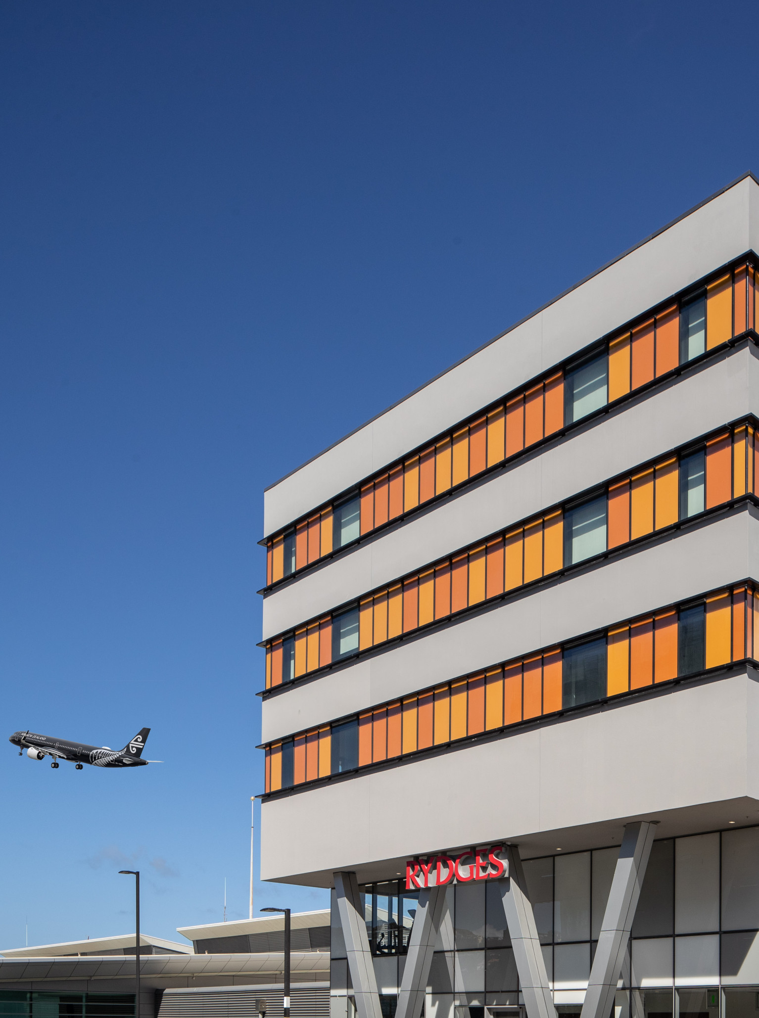 The modern Rydges Wellington Airport hotel features a striking grey and orange-paneled exterior with a plane against a clear blue sky.