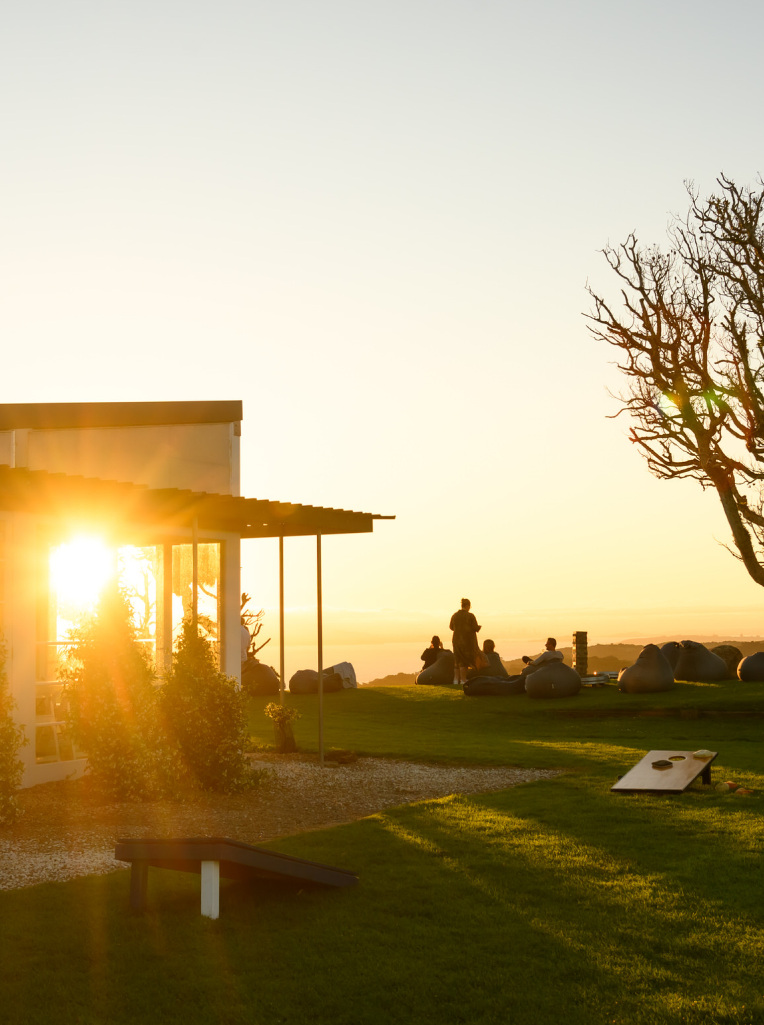 Guests relax on the lawn at Batch Winery, a modern Waiheke Island venue, with sunset views from its glass-fronted building.