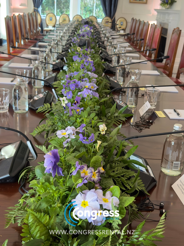 A formal conference room in a historic Wellington government venue, with AV by Congress Rental New Zealand, features a long table and a vibrant floral centerpiece.