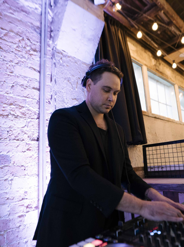 DJ Jarom Hall mixing music in an industrial-chic Auckland venue, featuring exposed brick and warm string lights.