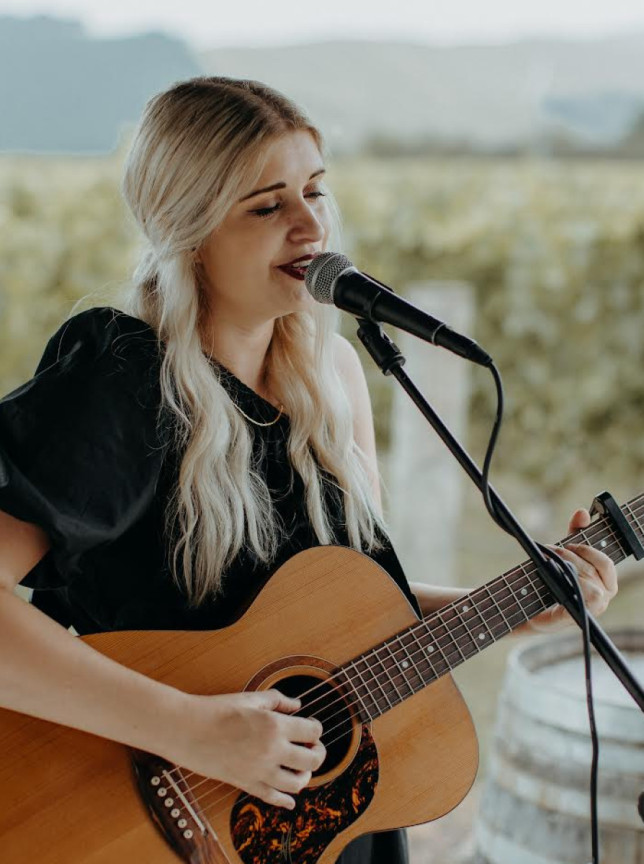 Rachel Hamilton, an Auckland-based acoustic musician, performs outdoors with her guitar and microphone, set against a picturesque vineyard backdrop.