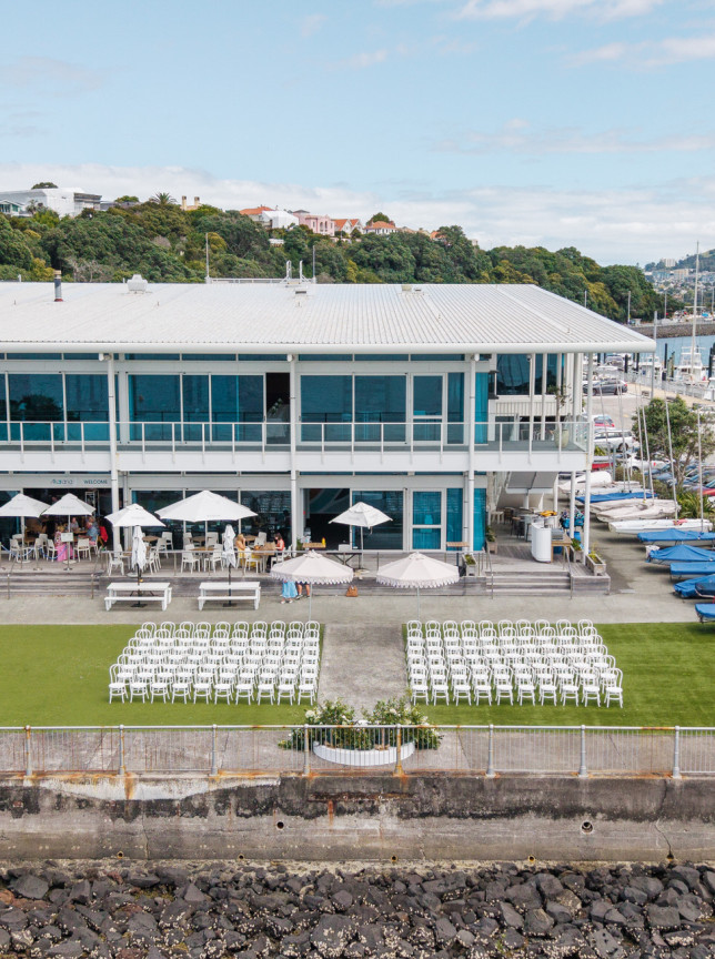 An outdoor wedding ceremony on the lawn of the modern waterfront Akarana Event Centre in Auckland, with a patio dining area overlooking the harbour.