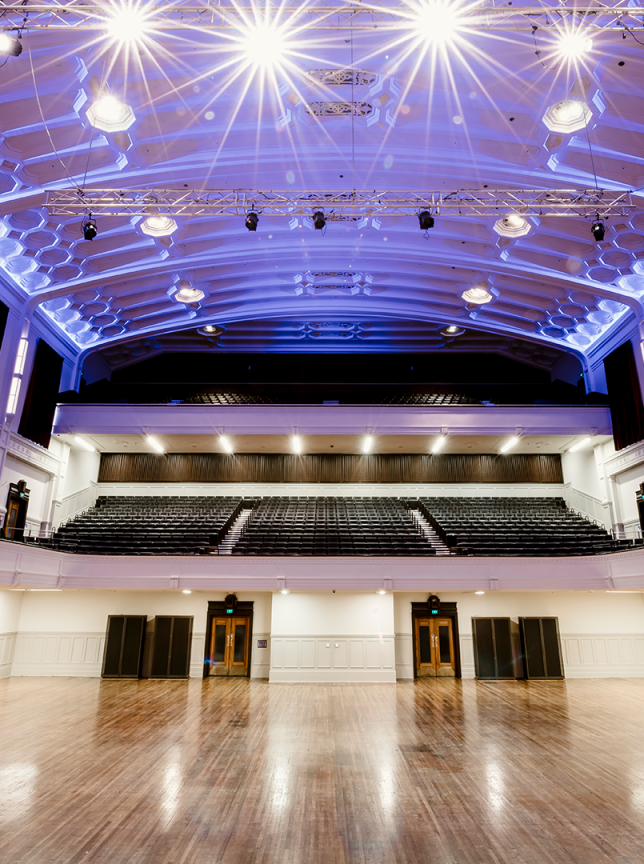 The Dunedin Centre's grand baroque main auditorium in Dunedin showcases tiered seating, a polished wooden floor, and striking blue lighting.