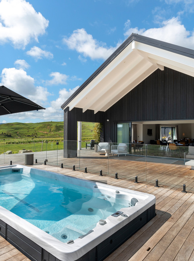 The modern Te Karaka Lodge in Waikato features an outdoor heated spa pool on a wooden deck, overlooking rolling hills.