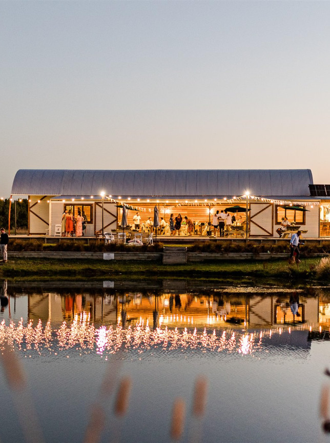 An evening event unfolds at Backpaddock Lakes, Hawke's Bay, filling its rustic barn venue with light that shimmers on the serene lake.