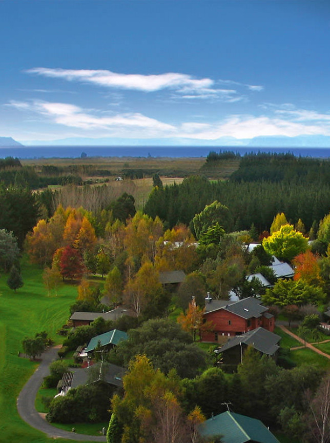Aerial view of Tongariro Lodge's rustic chalets in Taupo, an iconic fishing lodge, surrounded by autumn trees, river, and Lake Taupo.