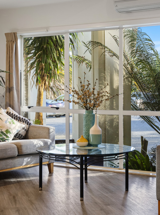 A modern living room at Taipa Beach Resort, Northland, features light grey sofas and tropical palm tree views.
