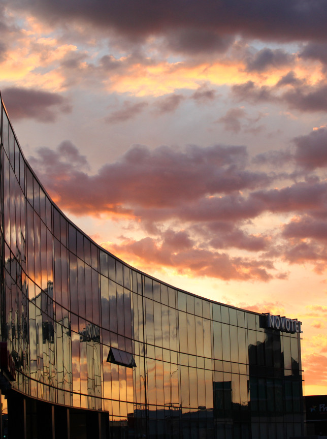 The modern glass facade of the Novotel New Plymouth Taranaki hotel reflects a vibrant sunset sky.