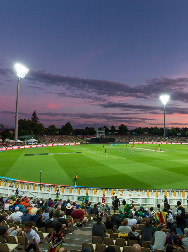Seddon Park in Hamilton presents a lively cricket match under floodlights at dusk, within its distinctive village green setting with tree-lined banks.