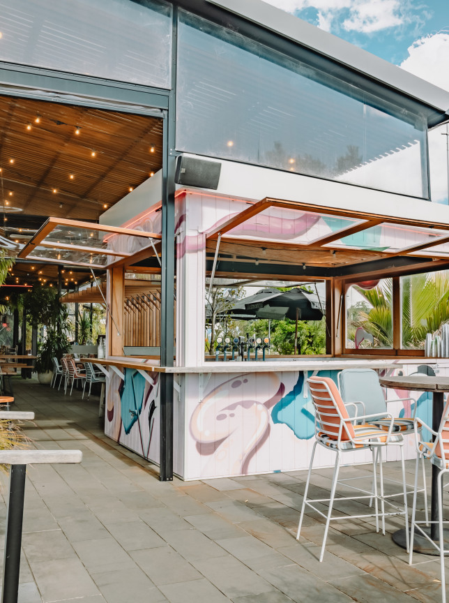 The modern, cabana-style outdoor bar area at Long Bay Surf Club in Auckland offers a relaxed beachside vibe.