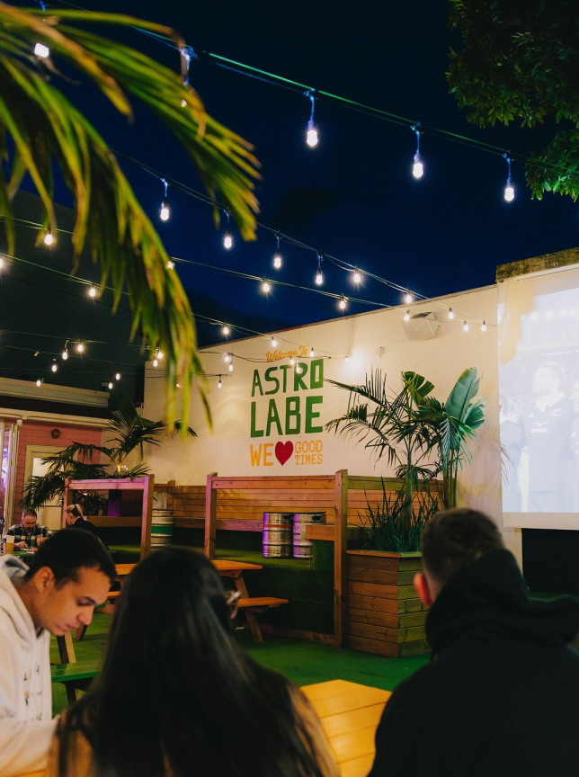 Astrolabe Brew Bar's vibrant Mount Maunganui outdoor beer garden, lit by string lights, with palms and a large projection screen.