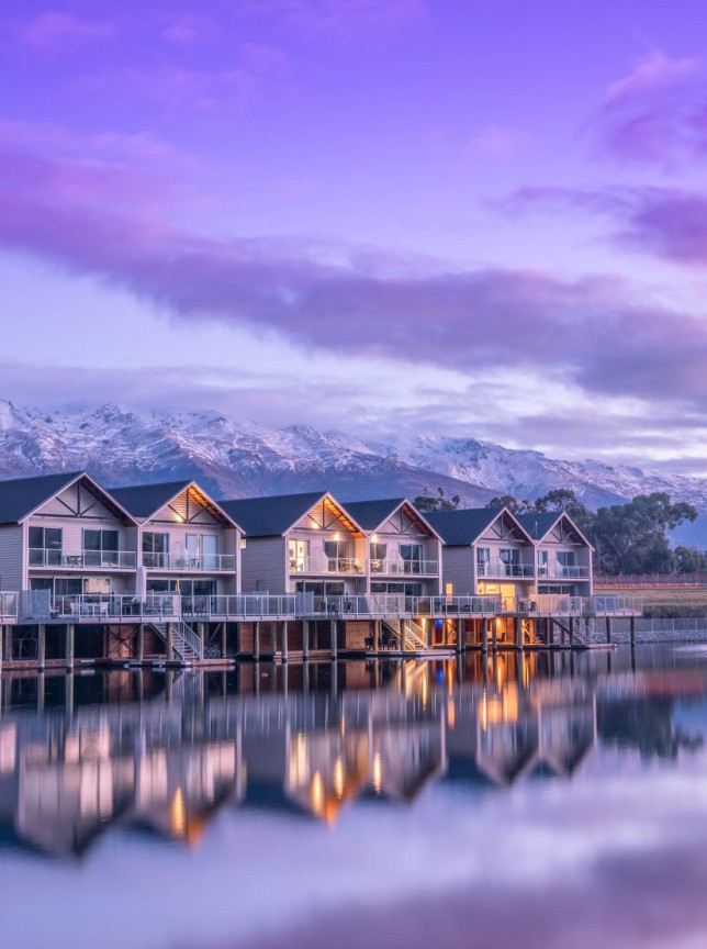 Boatshed-themed villas at Marsden Lake Resort, Central Otago, reflect on Lake Dunstan with snow-capped mountains under a colorful sky.