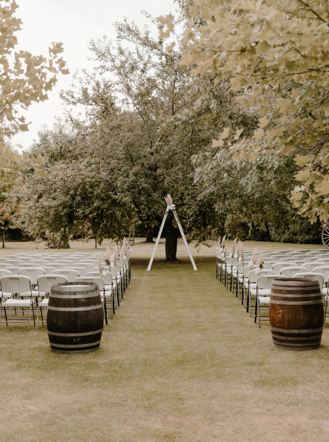 An outdoor wedding ceremony setup featuring white chairs, barrels, and a rustic arch within Omarino Estate's mature country gardens in Christchurch.