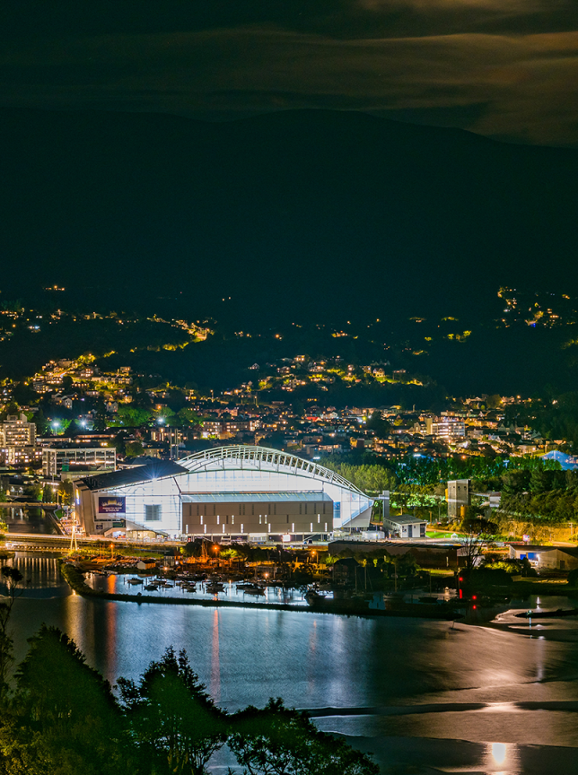 Dunedin's Forsyth Barr Stadium, a unique transparent-roofed "Glasshouse" venue, glowing at night amidst city lights.