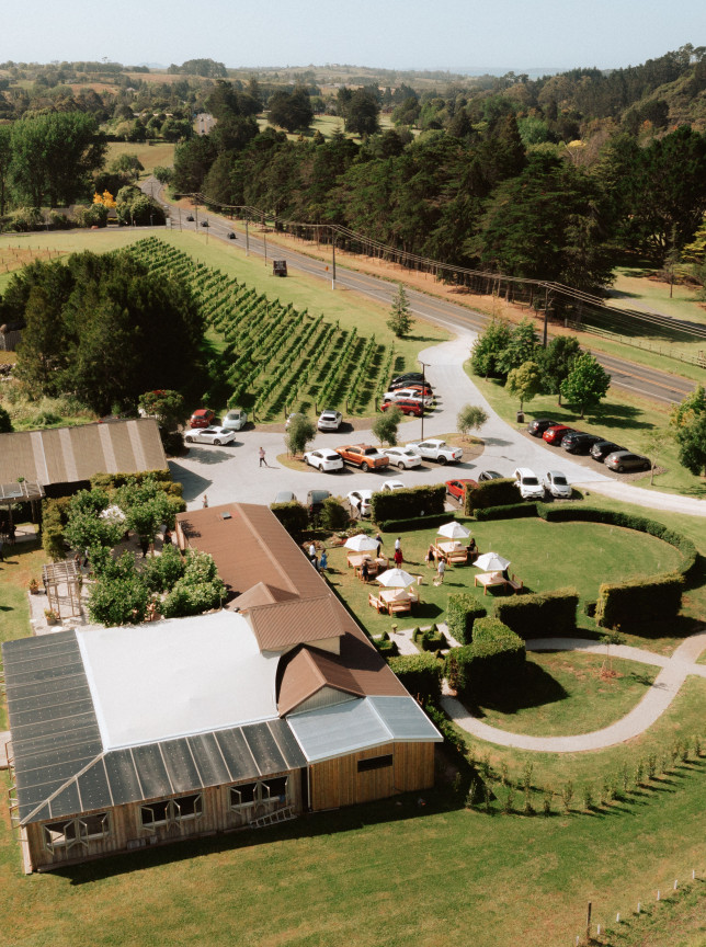 An aerial view of Turanga Creek Vineyard in Auckland, featuring rustic-modern buildings, an outdoor event space, and sprawling vineyards.