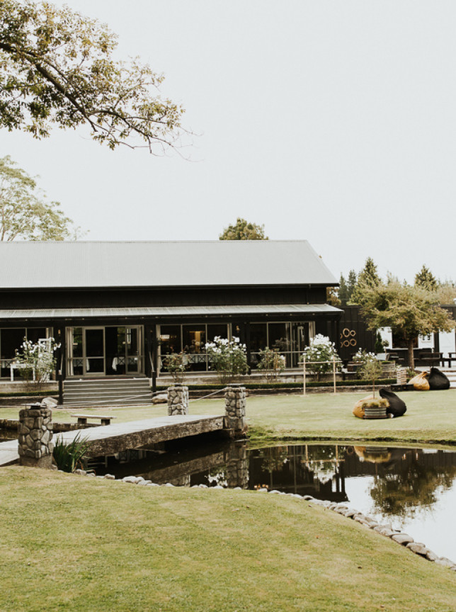 The modern event lodge at Stonebridge, Canterbury, is nestled among manicured gardens, featuring a tranquil pond and stone bridge.