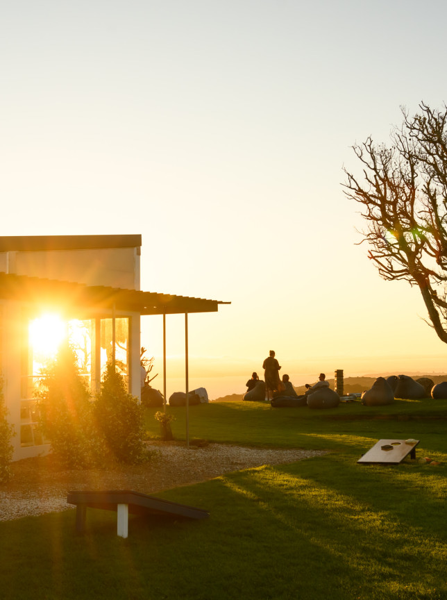 Guests relax on the lawn at Batch Winery, a modern Waiheke Island venue, with sunset views from its glass-fronted building.
