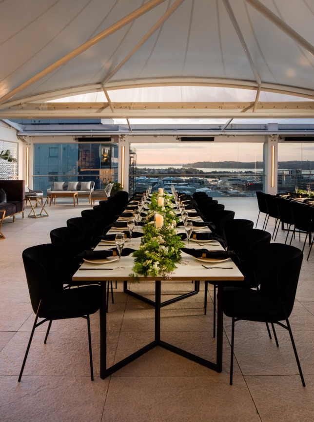 Rydges Auckland's modern rooftop event space showcases a long dining table under a retractable roof with panoramic city views.