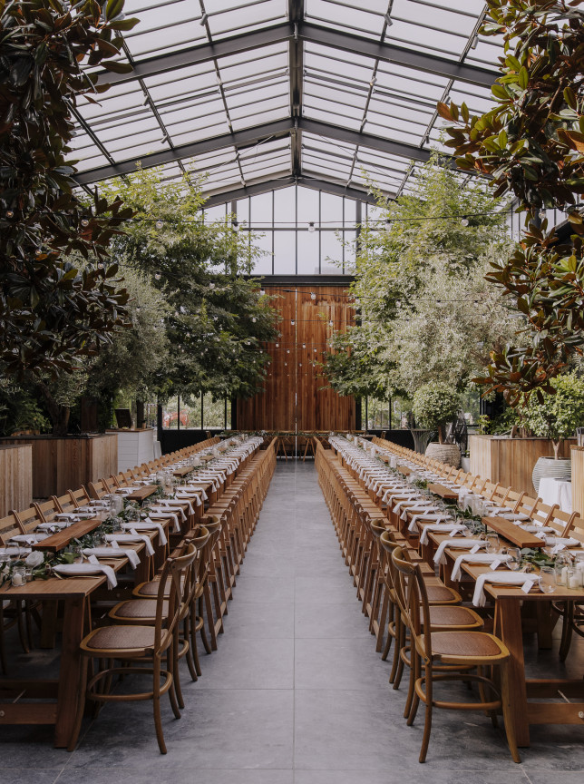 Long dining tables in the lush, glass-walled sanctuary of Glasshouse Morningside, Auckland, set for an elegant event.