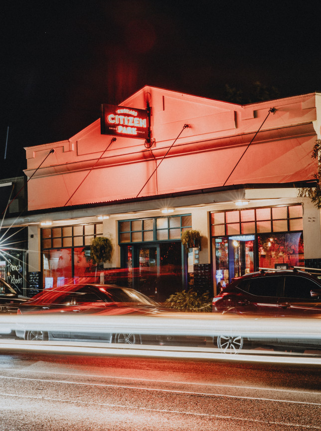 Citizen Park, an industrial-chic bar in Auckland, with its heritage facade and neon sign, illuminated at night by car trails.