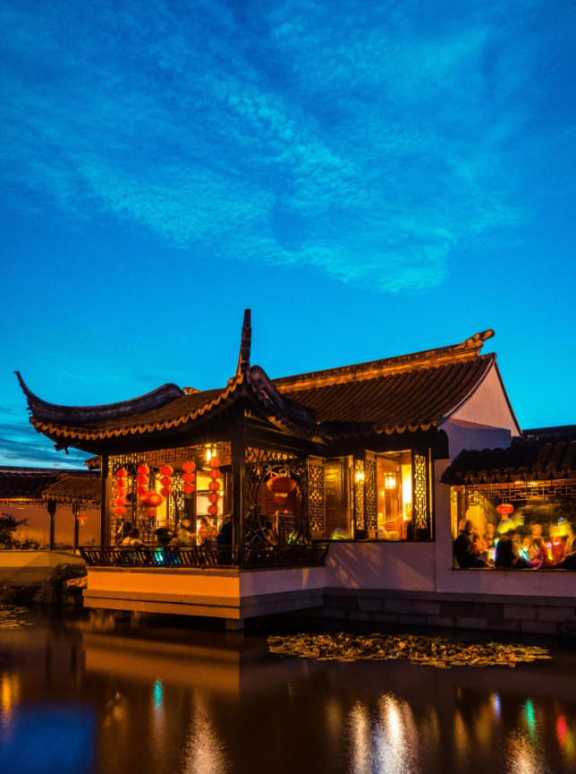 An illuminated traditional Chinese pavilion in Dunedin Chinese Garden, designed in authentic Jiangnan style, welcoming guests by the water.