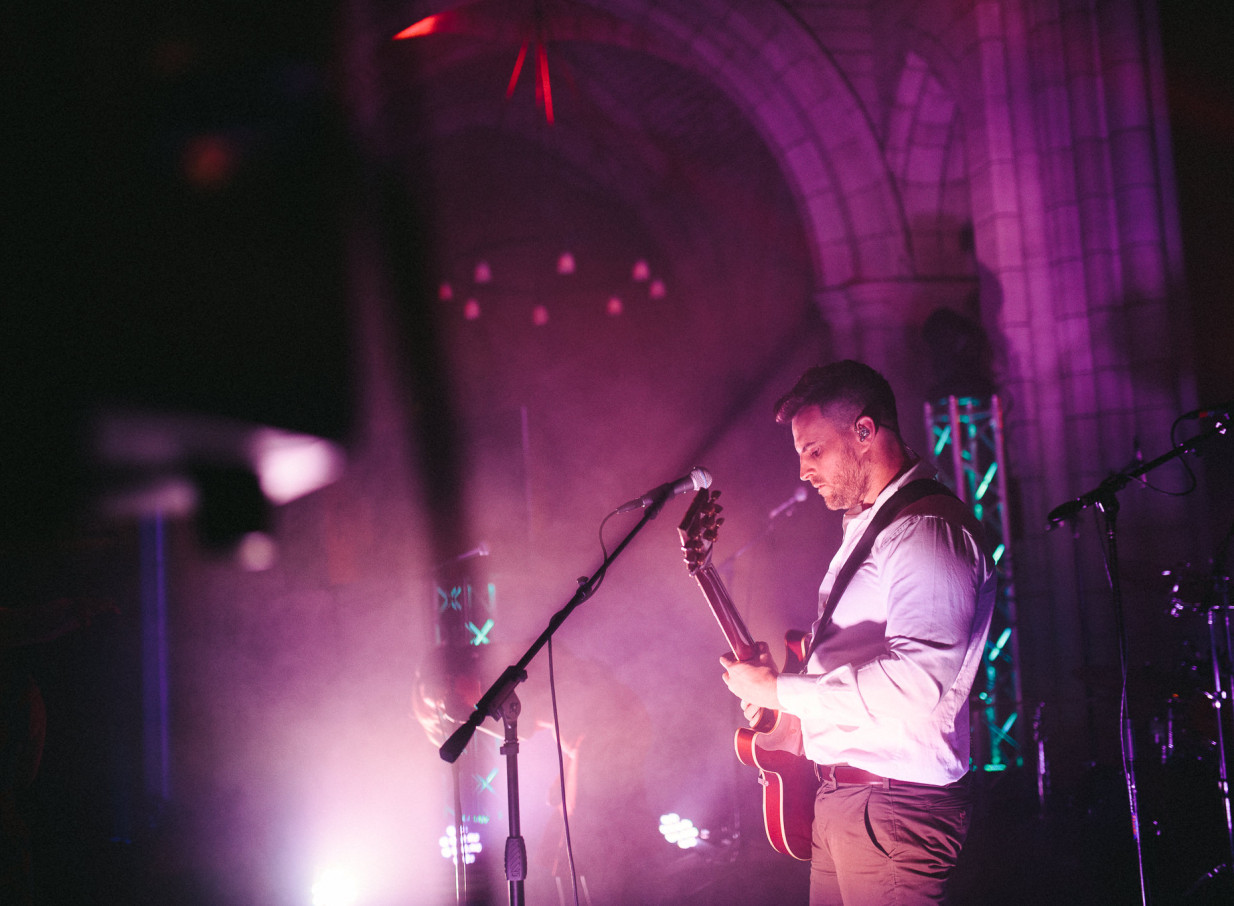 A Blue Steel guitarist plays on stage with purple lighting, highlighting the Imperial Baroque architecture of Auckland Town Hall.