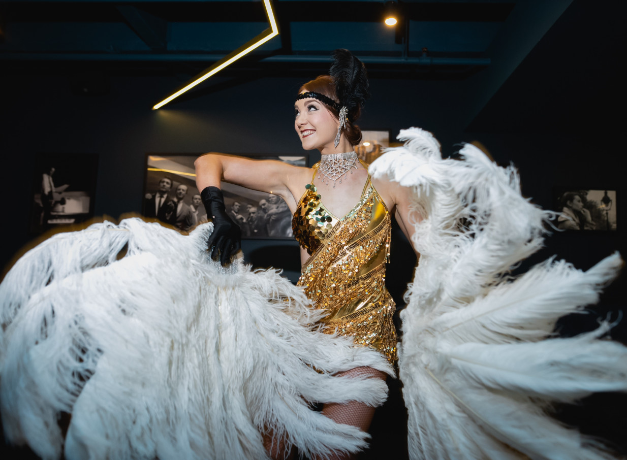 An energetic Finesse Entertainment showgirl, based in Auckland, dazzles in a gold sequin costume with large white feather fans.
