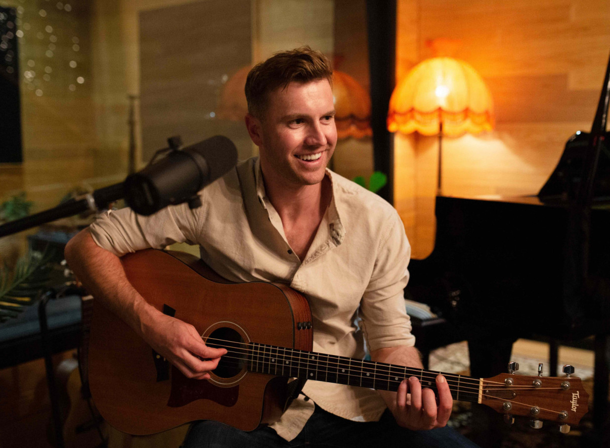 Auckland acoustic musician Daniel John, known for his easy-listening style, plays guitar in a warm studio setting.