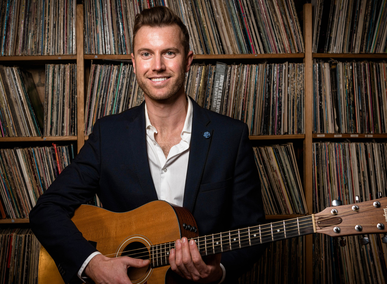 Auckland acoustic musician Daniel John poses with his guitar against a wall of vintage vinyl records.