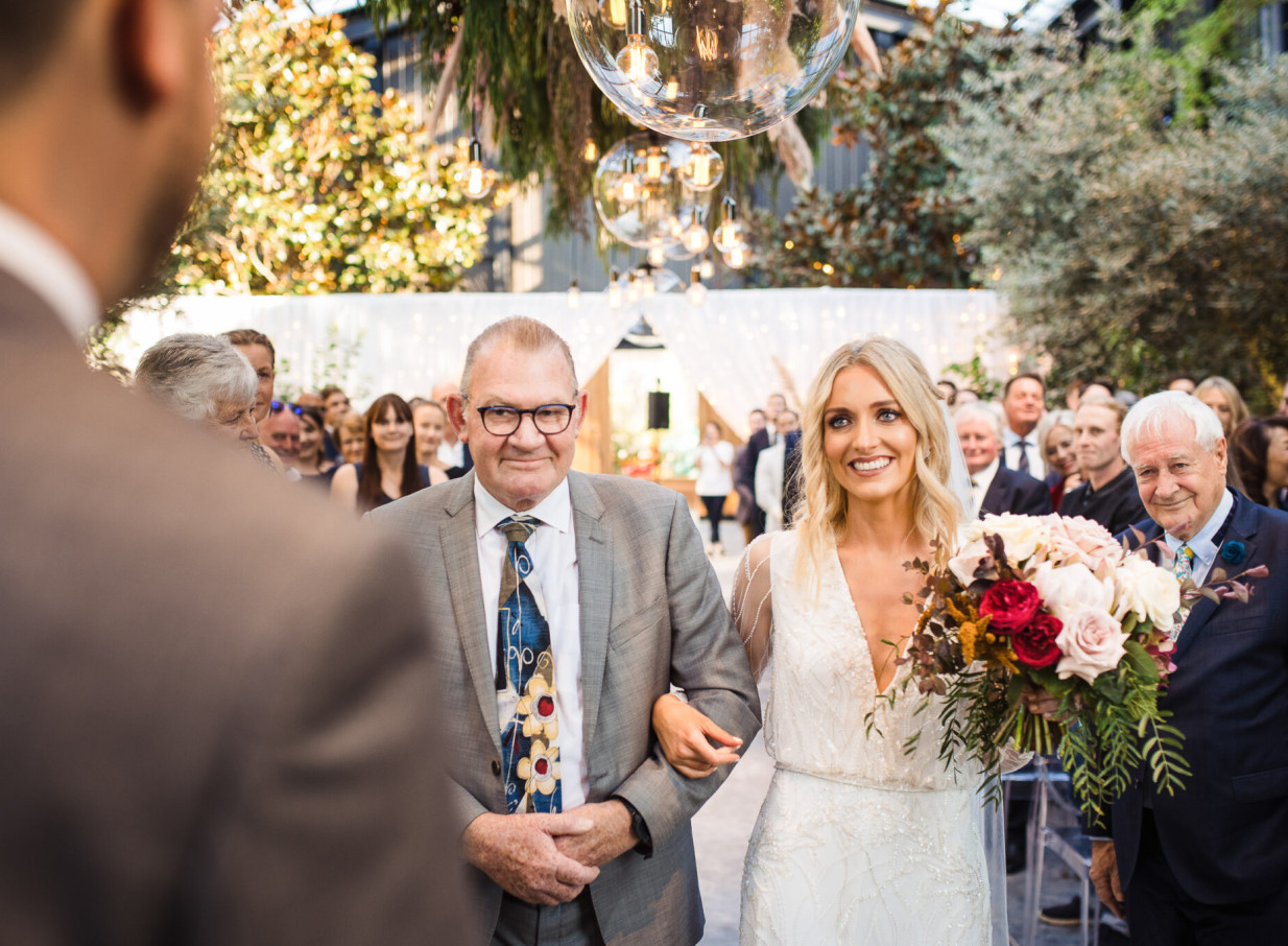 A bride and her father walk down the aisle in an elegant greenhouse wedding ceremony, captured by Zahn in Auckland.