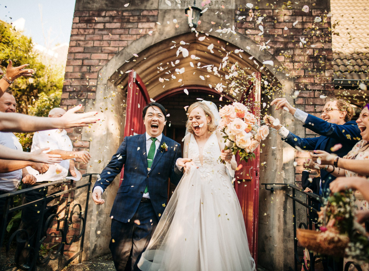 A joyous wedding couple exits a historic brick building in Auckland, showered with confetti.