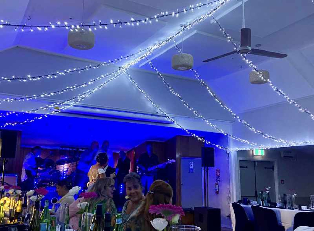 An indoor event space in Auckland, styled by Light and Leaf Co., features draped fairy lights and blue uplighting over a band and seated guests.