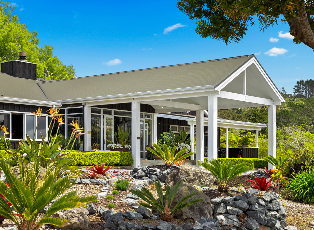 The contemporary lodge exterior with a covered entrance and lush tropical gardens welcomes guests to Woodhouse Mountain Lodge, Auckland.