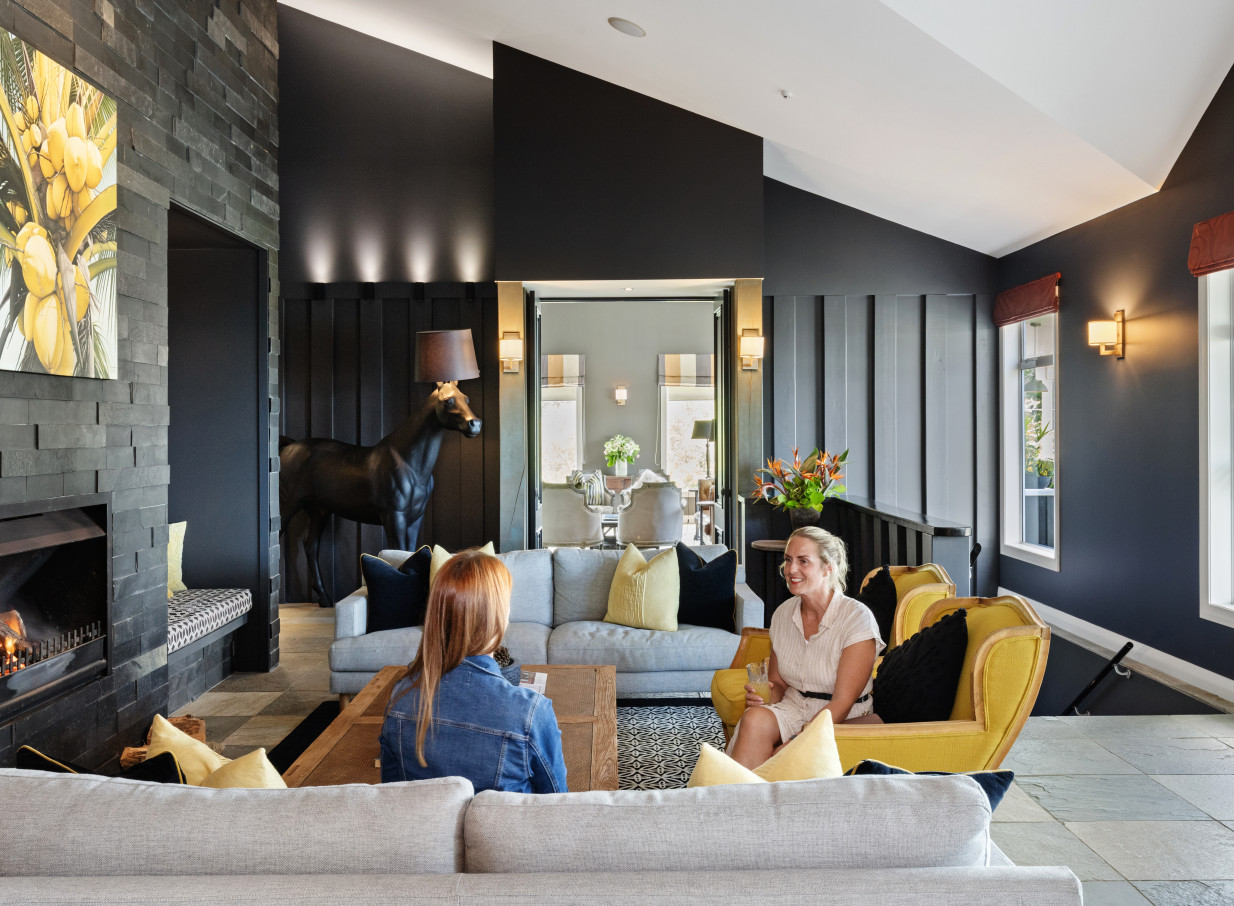 The contemporary lodge lounge at Woodhouse Mountain Lodge in Auckland features two women conversing by a cozy fireplace.