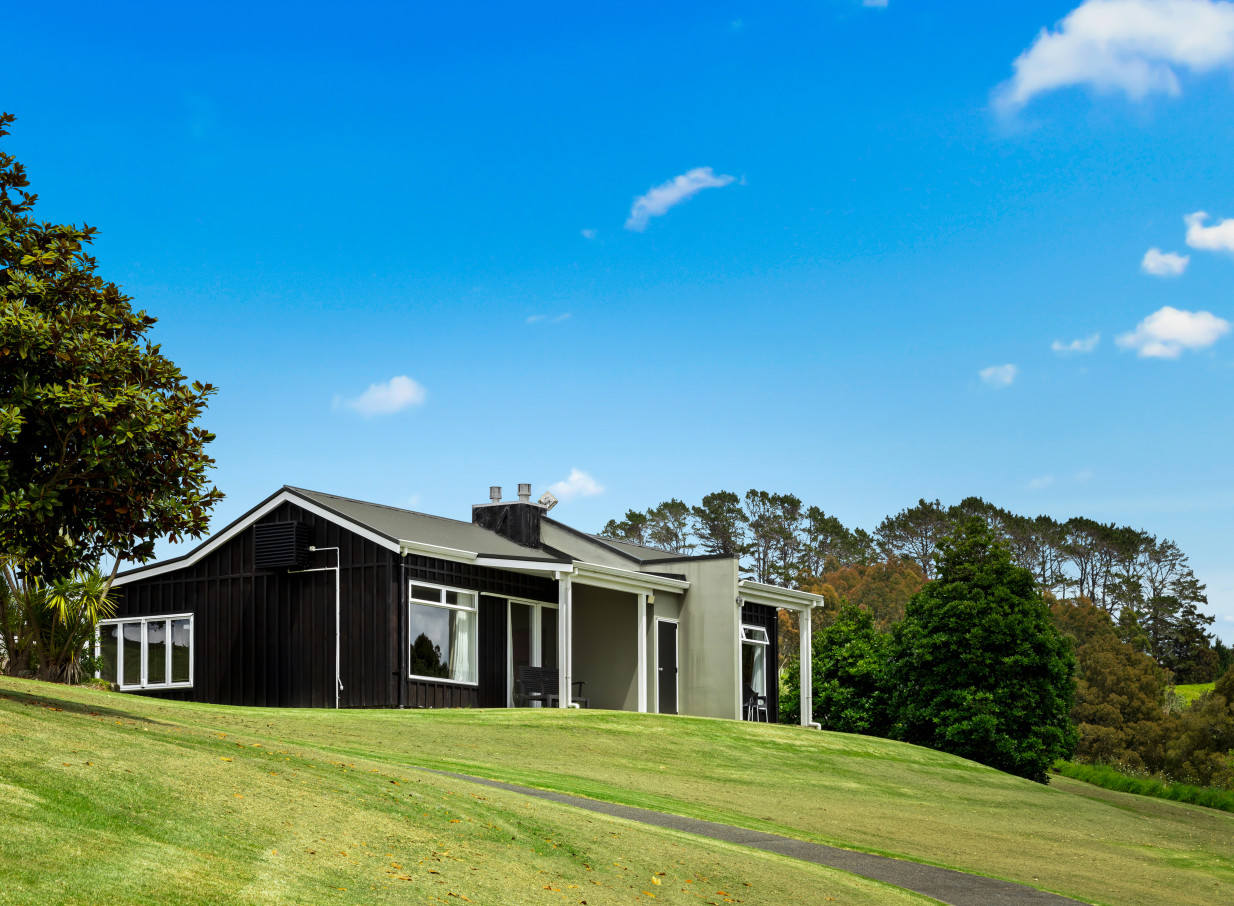 A contemporary villa at Woodhouse Mountain Lodge, Auckland, set on a bright green, grassy hillside under a clear blue sky.