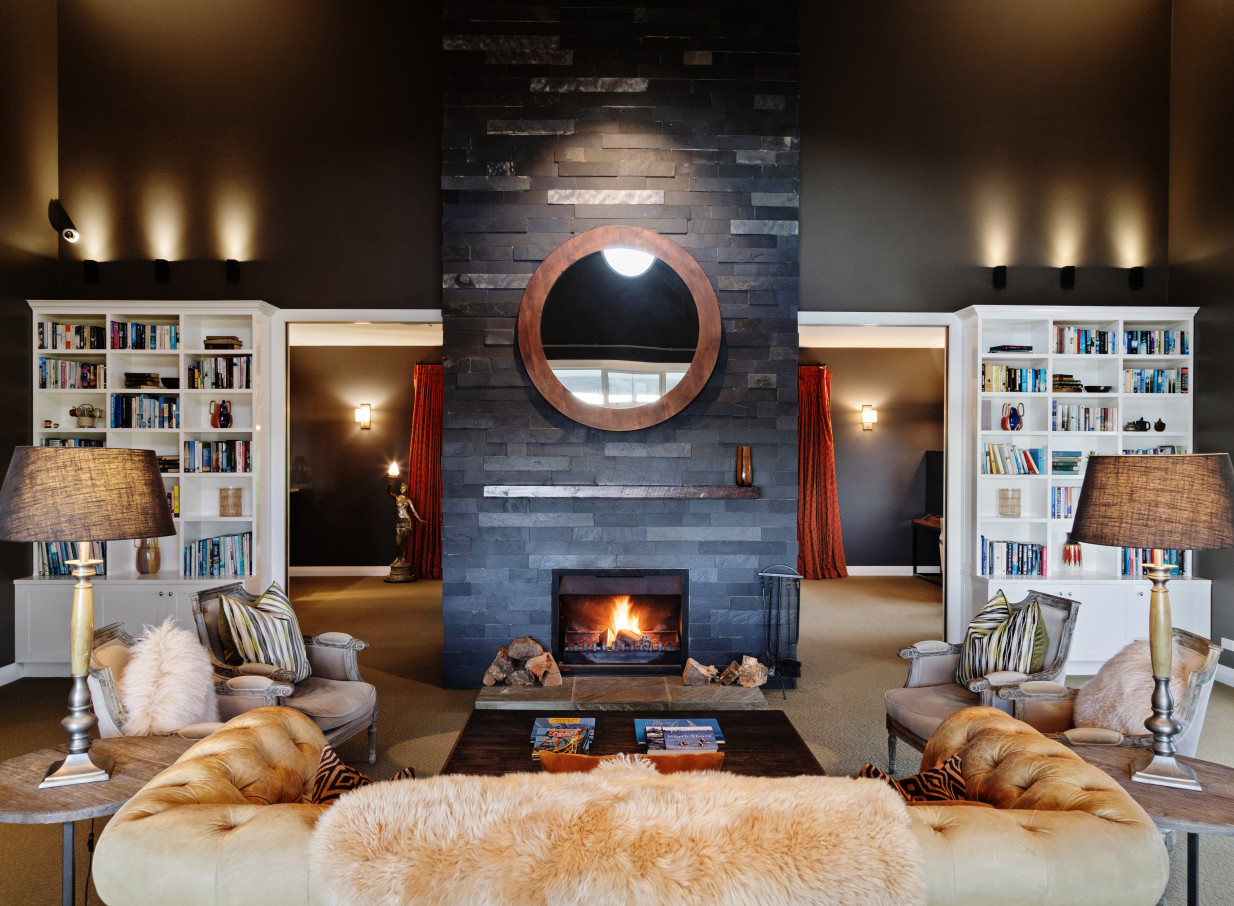 A luxurious mountain lodge library lounge at Woodhouse Mountain Lodge, Auckland, with a cozy stone fireplace and plush seating.