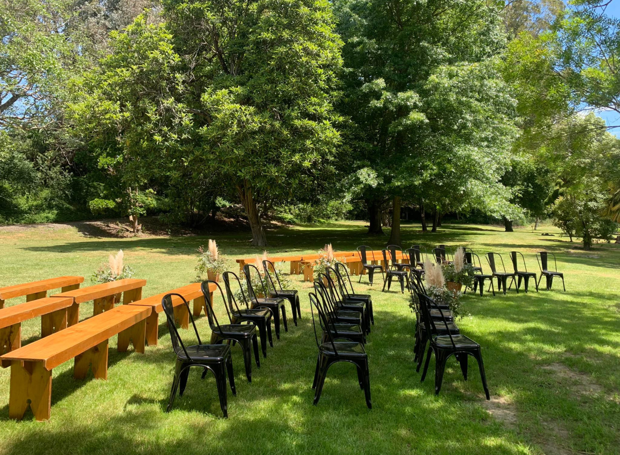 An outdoor ceremony on a green lawn with wooden benches and black chairs at Omatua, a natural garden venue in Hawke's Bay.