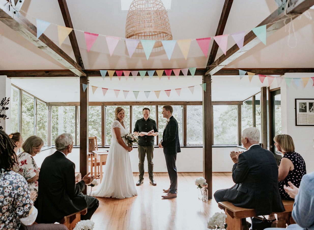 An indoor wedding ceremony in the rustic lodge-style venue Omatua, nestled in Hawke's Bay, featuring wooden beams and large windows.