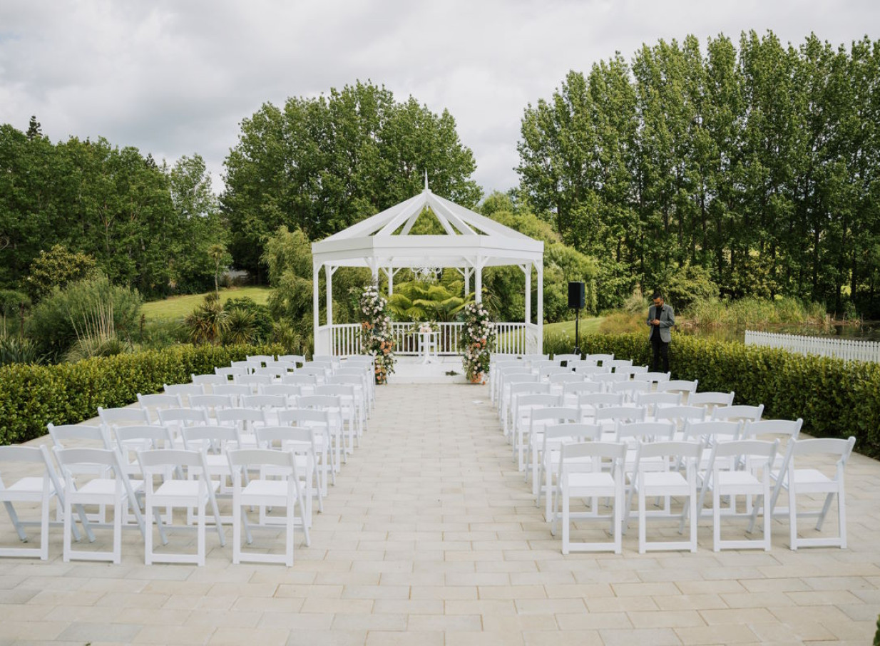 An elegant outdoor garden ceremony at Abel Estate in Auckland, featuring a modern white rotunda and rows of white chairs.