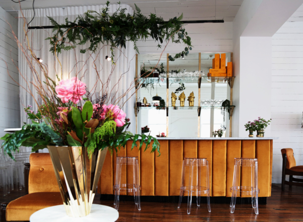 A chic, elegant bar space at The Canapé Room, Wellington, with a gold velvet front, clear stools, and lush floral decor.