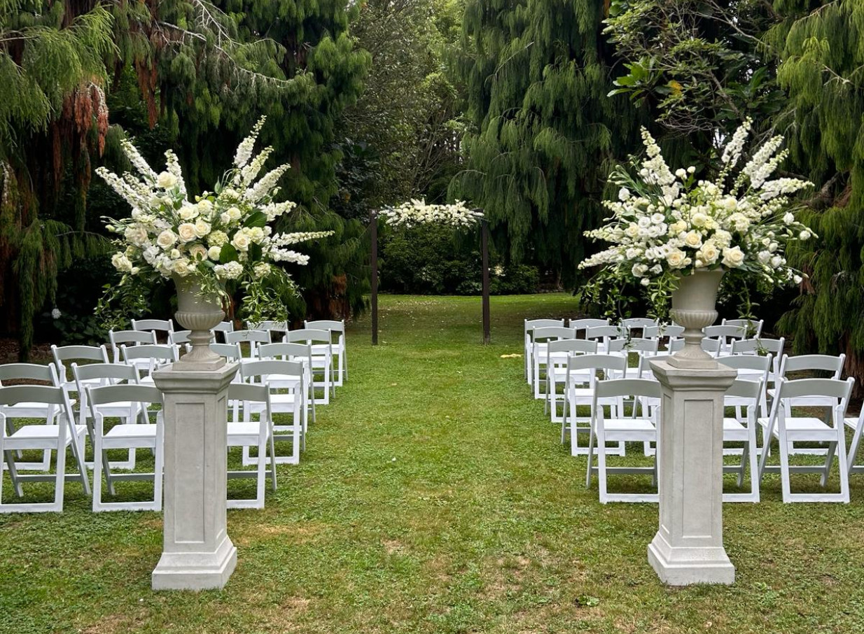 An outdoor ceremony setup featuring white chairs and floral arrangements in the botanic garden setting of Anam Cara Gardens, Wellington.