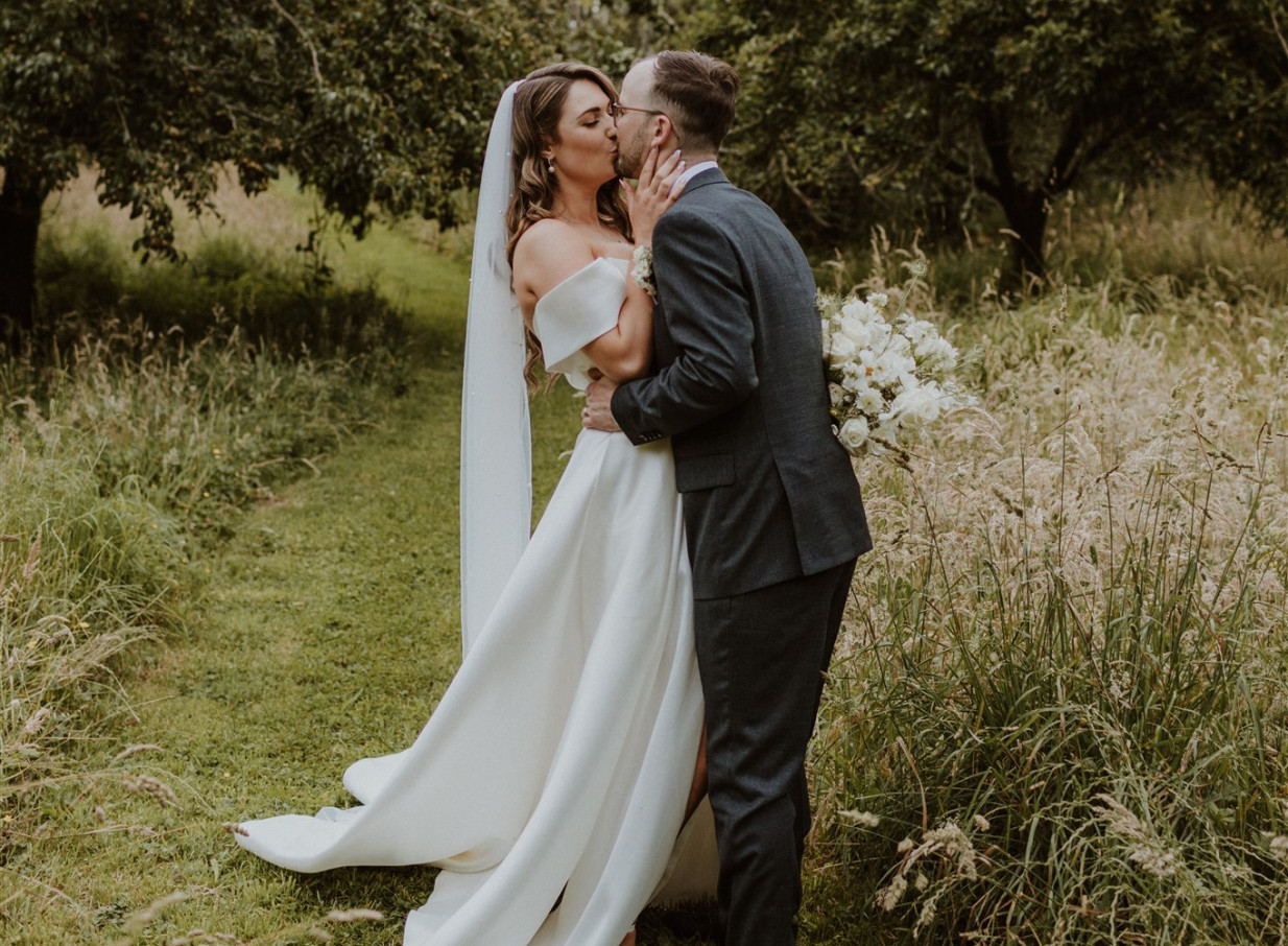 A couple shares a romantic outdoor wedding kiss in the picturesque garden setting of Anam Cara Gardens, Wellington.